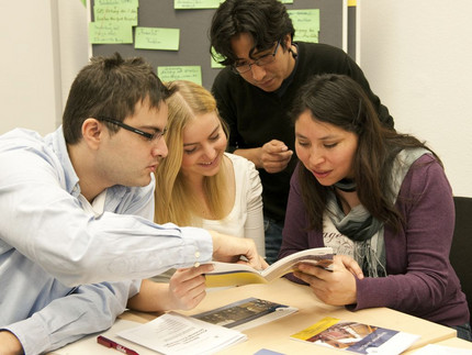 Das Bild zeigt eine Gruppe Studenten. Sie schauen zusammen in ein Heft. Ein Mann zeigt mit dem Finger auf etwas in dem Heft.