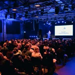 Bühne in einer Halle, darauf ein Redner, davor Publikum auf Stühlen