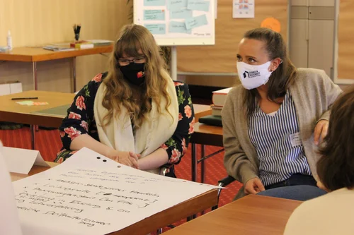 Das Bild zeigt zwei Frauen mit Mund-Nasen-Schutz, die an einem Tisch in einem Seminarraum sitzen. Vor ihnen liegt ein großes Blatt Papier mit handschriftlichen Notizen. Beide wirken in ein Gespräch vertieft. Im Hintergrund sind Tische, Moderationsmaterial und Pinnwände zu sehen.