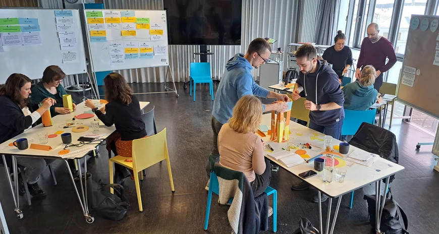 Verschiedene Menschen sitzen an Tischen im Rahmen eines Workshopsettings und haben viele verschiedene Materialien auf dem Tisch.
