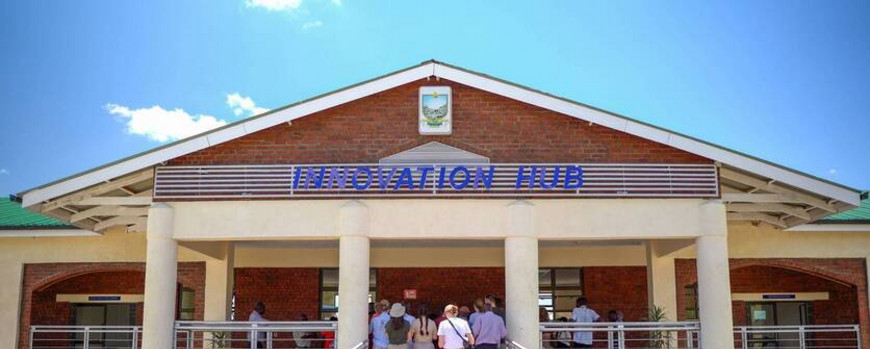 Auf dem Foto ist ein modernes Gebäude mit spitzem Dach zu sehen, welches mit einem Wappen und dem Schriftzug "Innovation Hub" über dem Eingang verziert ist. Ein langer Weg mit Geländer führt auf den Eingang zu und vor dem Eingang stehen Menschen schwarzer und weißer Hautfarbe gemeinsam mit dem Rücken zur Kamera.