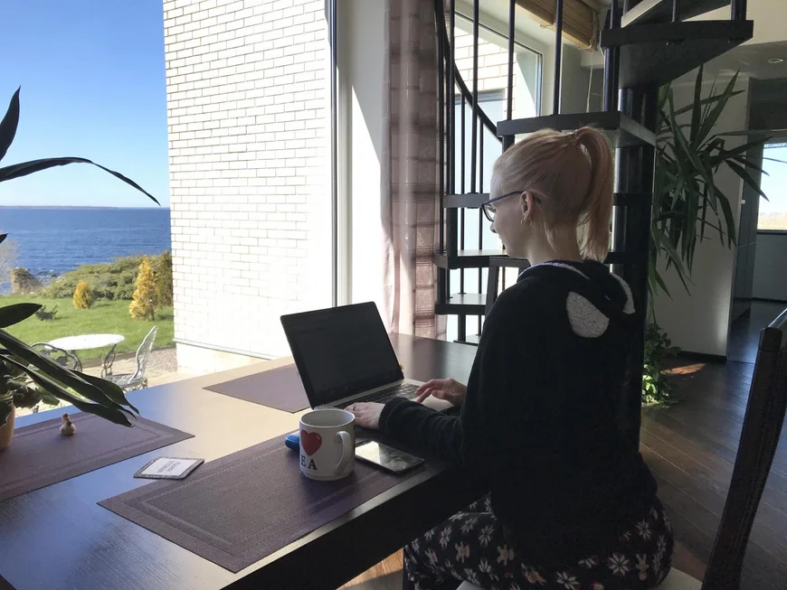 ein stidentin sitzt auf einer Terasse mit Laptop an einem tisch. SCheint ein imposantes Haus zu sein, im Hintergrund sieht man das Meer, man gehts uns schlecht heute.