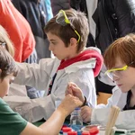 Zwei Kinder mit Kitteln und Schutzbrillen führen ein Experiment durch