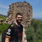 Eine Person im Vordergrund lacht in die Kamera. Hinter ihr Ruinen und grüne Vegetation. Die Person und die Ruine befinden sich auf einem Berg.