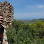Eine Person im Vordergrund lacht in die Kamera. Hinter ihr Ruinen und grüne Vegetation. Die Person und die Ruine befinden sich auf einem Berg.