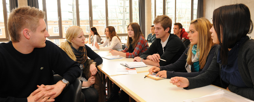 Das Bild zeigt eine Gruppe von Student:innen in einem Seminarraum. Sechs der Student:innen sitzen als Gruppe zusammen und schauen sich an.