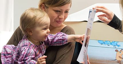 Kind auf dem Arm der Mutter, zeigt auf eine Urkunde, die ihr hingehalten wird