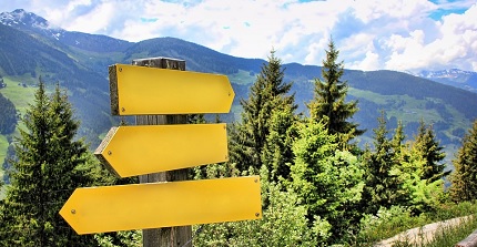 Ein Wegweiser mit gelben Schildern, dahinter Nadelbäume und am Horizont Berge.